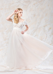 fashionable wedding dress, beautiful blonde, bride hairstyle and makeup concept - posing young pretty woman in long luxury white gown indoors on light background, romantic female model in a studio
