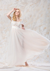 fashionable wedding dress, beautiful blonde, bride hairstyle and makeup concept - posing young woman in long luxury white gown indoors on light background, smiling female model in a studio