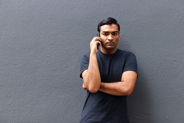 Handsome man wearing black shirt standing against gray wall with arm crossed talking on phone angry