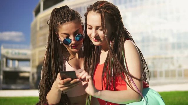 Happy women with dreads sitting on grass in summer park and using a smartphone. Young friends talking and watching photos, surfing in the internet outdoors having fun. Slowmotion shot