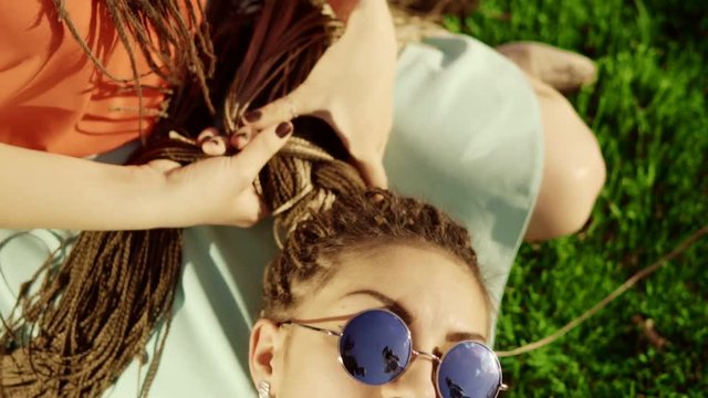 Young hipster girls relaxing on the grass in park. One girl is weaving braids for another. two hipster women laying on the grass, enjoying nice day Best friends girls having fun, joy