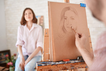 Obraz premium Portrait of artist drawing portrait of young woman sitting in front of him in art studio, focus on the picture in pencil