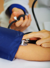 Close up of a young doctor using the stethoscope to hear the pulse while a woman is with a tensiometer in her arm, in a doctor consulting room background