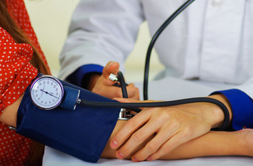 Close up of a young doctor using the stethoscope to hear the pulse while a woman is with a tensiometer in her arm, in a doctor consulting room background