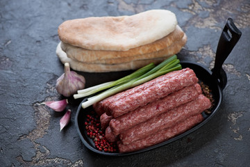 Cevapi or cevapcici sausages in a frying pan with green onion and pitas. Studio shot over brown stone surface