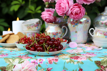 Ripe cherries, red currants and cookies in a vase. A bouquet of nostalgic pink roses. Tea-set. Table setting and blue tablecloth with roses. Romantic dinner in the garden by candlelight.