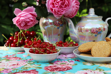 Ripe cherries, red currants and cookies in a vase. A bouquet of nostalgic pink roses. Tea-set. Table setting and blue tablecloth with roses. Romantic dinner in the garden.