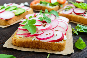 Several kinds of sandwiches with vegetables: radish, tomatoes, cucumber, arugula on crispy toast on a dark wooden background. Vegetarian dish. Proper nutrition. Healthy food. Breakfast.