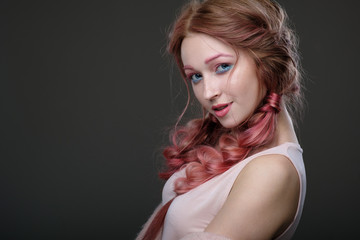 Fototapeta premium Close-up portrait of girl with pink hair in braids, pink and blue makeup, standing in a half-circle on a dark background playfully looking into the camera