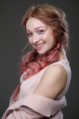 Fototapeta premium Close-up portrait of girl with pink hair in braids, and pink-blue makeup with a pink coat worn off the shoulder standing in the half-turn on a dark background smiling and looking at camera