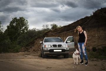 Brutal, daring girl with a fighting dog breed of bull terrier, near his car against the background of the cliff in the evening