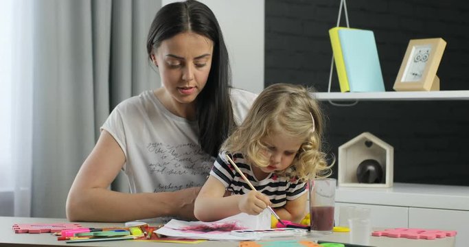 Charming little girl with blond curly hair and her mother with long black hair driwing using paints, looking on camera and smiling with teeth. Indoor.