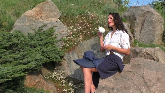 Happy woman is drinking tea sitting on a rock in the park.