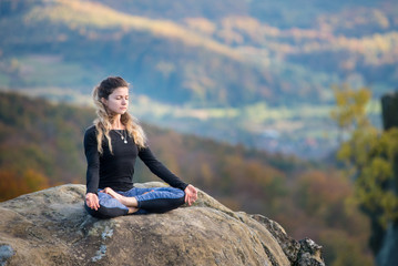 Naklejka na ściany i meble Happy slim girl is practicing yoga and doing asana Siddhasana on the top of the mountain. Autumn forests, rocks and hills on the background