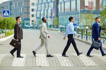Multi-ethnic group of confident coworkers crossing road in city center, modern office buildings on...