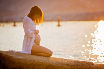 Beautiful pregnant woman, sitting on the shore on the beach on sunrise