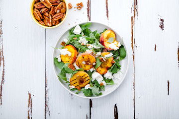 Fresh salad with grilled peach halves, arugula and burrata on a plate on white distressed wooden background. Top view. Summer food concept