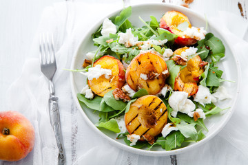 Fresh salad with grilled peach halves, arugula and burrata on a plate on white distressed wooden background. Top view. Summer food concept