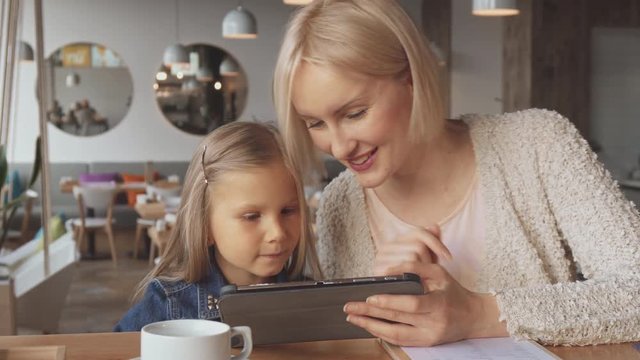 Cute young woman and her daughter using tablet at the cafe. Pretty blond woman telling something to her child. Attractive caucasian little girl pointing her finger on the screen