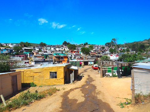 Street Of Colorful Informal Settlements (Slum), Huts Made Of Metal In The Township Or Cape Flats Of Stellenbosch, Cape Town, South Africa With Blue Sky And Clouds Background