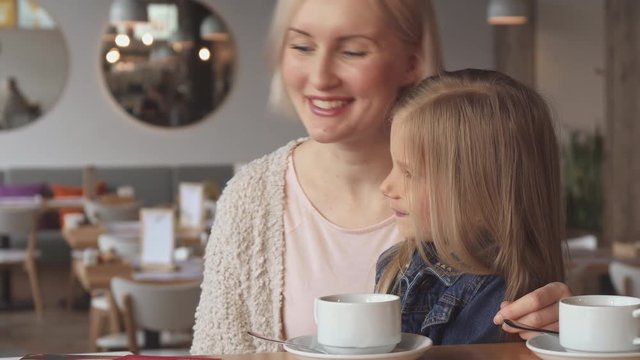 Close up of cute caucasian little girl whispering in her mother's ear at the cafe. Pretty blond woman listening what her daughter saying. Attractive female child sitting in her mom's lap