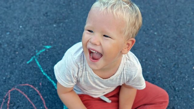 happy boy sitting on asphalt with chalk paintings smiling on camera slow motion