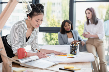 Smiling young asian businesswoman working with blueprint while colleagues sitting behind