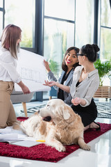 smiling multiethnic businesswomen looking at blueprint with dog at office