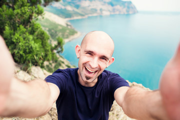 Young man taking travel selfie on trekking excursion day - Hipster guy self photo at view point with blue ocean background - Concept of healthy lifestyle in beauty of nature