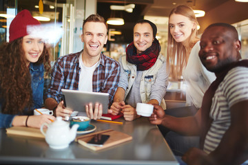 Group portrait of cheerful young businesspeople looking at camera with toothy smiles while brainstorming in lovely coffeehouse