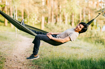 Happy peruvian young man resting and relaxing in hammock outdoor on nature in forest in summer sunny day with pine trees and green grass. Travel, holidays, tourism, vacation. Dreaming in park.