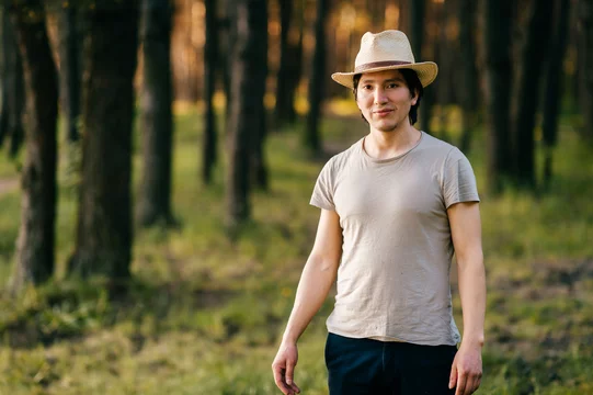 Young and lonely native american indian latino indigenous man standing in forest outdoor in summer in straw hat, casual clothing above bonfire with smoke on vacation. Evening sunset. Weekend.