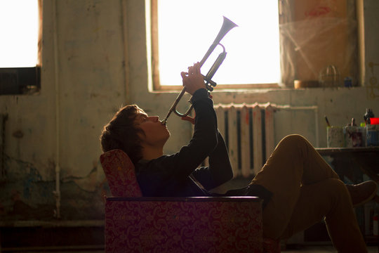 Young man sitting on vintage armchair in artist studio playing trumpet