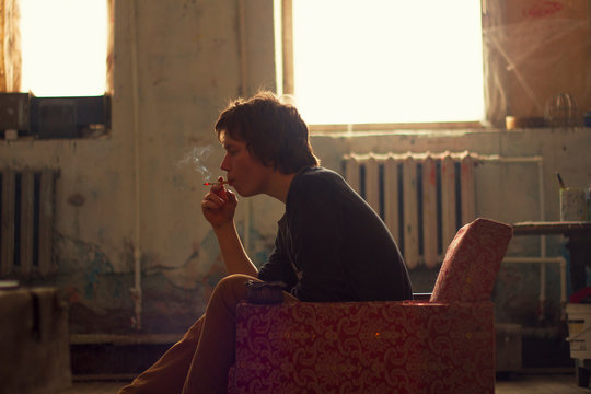 Young man sitting on vintage armchair in artist studio smoking cigarette