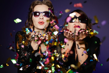 Having fun with best friend: two stylish women standing against dark background and blowing...