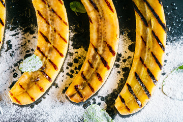 Shale stone plate with grilled bananas dessert decorated with sugar powder and green mint leaves. Macro Closeup View from Above.