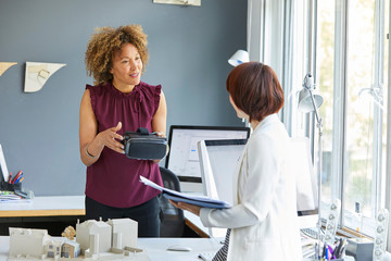 Female architect explaining virtual reality headset to colleague at office desk