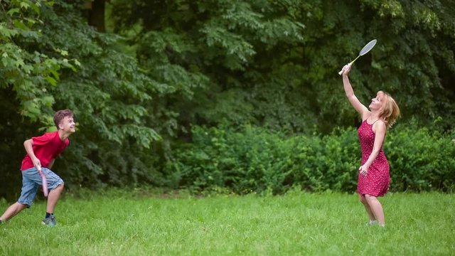 Mother and son playing badminton in meadow with forest in background. Child with badminton rackets in hands. Kid have fun in summer park at day.