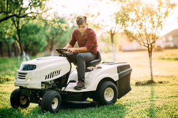 Ride-on lawnmower tractor with worker doing landscaping works at weekend sunset