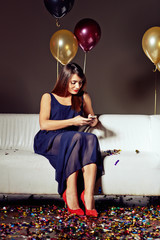 Full length portrait of attractive young woman texting with her friend on smartphone while sitting on sofa at night club