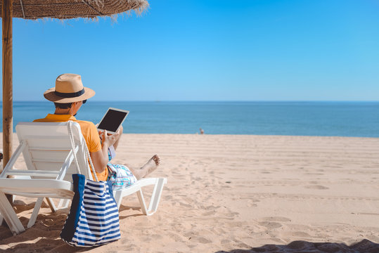 Back view of tourist using tablet pc on the beach. Holiday relaxation vacation photography on sunny blue sky seacoast, sea shore outdoors background
