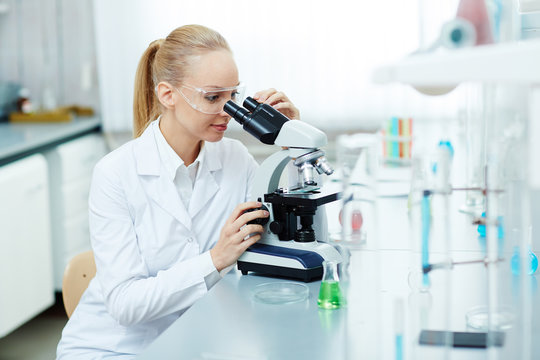 Portrait of beautiful woman using microscope working in modern laboratory, performing chemical tests and smiling