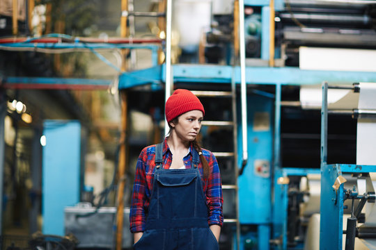 Confident young engineer wearing denim overall standing in warehouse with hands in pockets and looking away, waist-up portrait