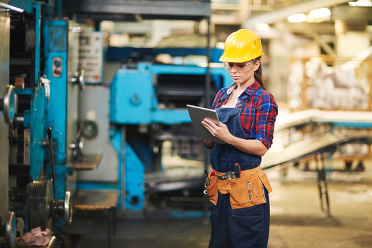 Portrait of pretty young technician wearing denim overall using digital tablet while carrying out inspection in production department of modern plant