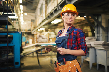 Waist-up portrait of pretty young engineer looking away while working on digital tablet,  interior of manufacturing plant on background