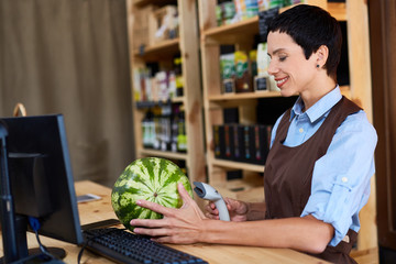 Profile view of beautiful middle-aged cashier scanning barcode of watermelon in small store with organic food