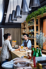 Profile view of young customer standing at cashdesk of small shop with organic products and talking to pretty middle-aged shop assistant
