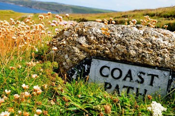 Coast path stone sign in the floral summer meadow. In the back are hills and sea (Cornwall, United Kingdom)