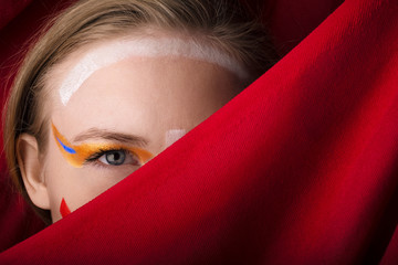 Girl with color make-up on a red background. A blonde with colored make-up peeks out because of the red cloth.