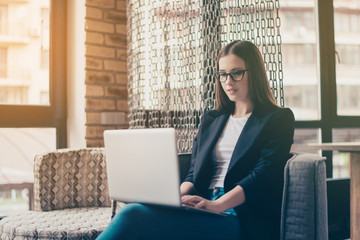 Serious young entrepreneur economist in formal black jacket, glasses, sitting at her work station...